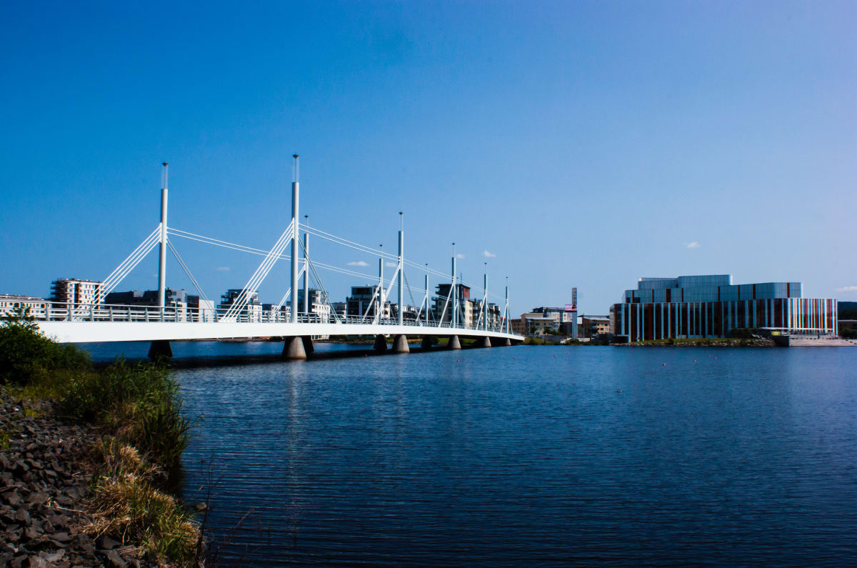 En modern vit gångbro sträcker sig över en vattenyta under en klarblå himmel. På andra sidan vattnet syns en modern byggnad med rödbruna och grå vertikala paneler, samt bebyggelse och grönskande växtlighet vid strandkanten.