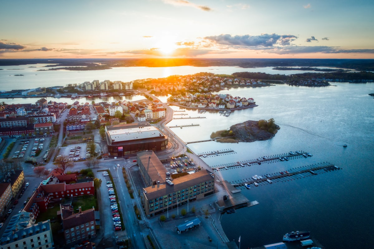 En vidvinkelbild tagen från ovan, visar en stadskärna vid vattnet vid soluppgång eller solnedgång. Flerbostadshus, småhus, parkeringsplatser, båthamnar med förtöjda båtar och broar syns i stadsmiljön. Vattenytan reflekterar solens ljus.