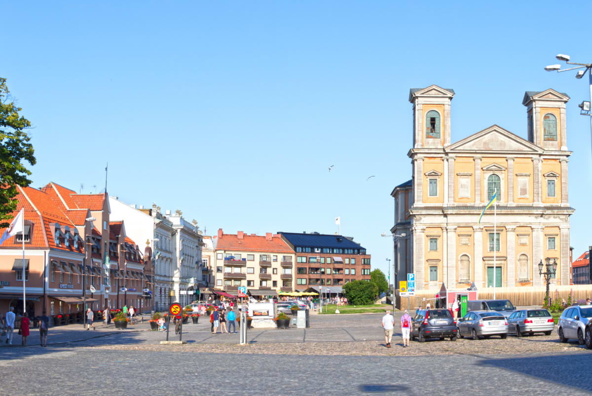 En stor, kullerstensbelagd torgplats omgiven av flera byggnader, inklusive en stor barockkyrka i orange och grått. Ett flertal personer promenerar på torget, och parkerade bilar syns till höger. Himlen är klarblå och inga moln syns.