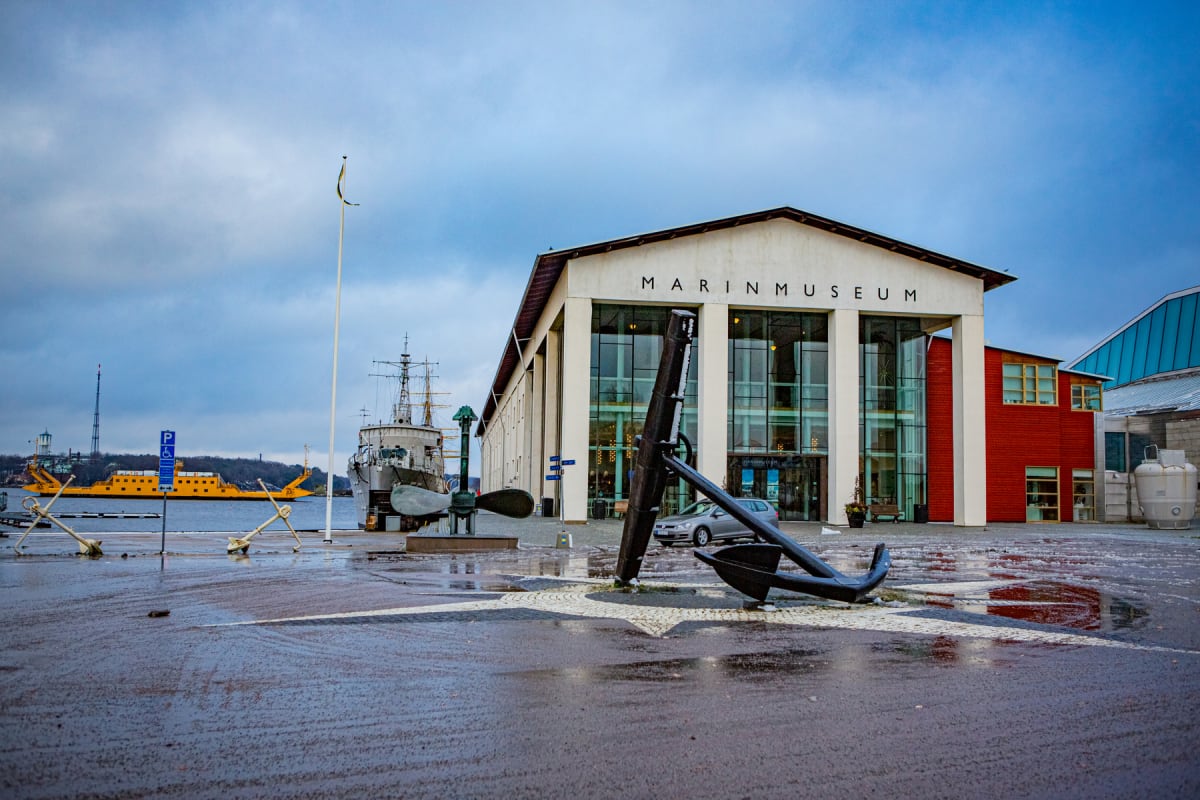 En exteriörbild av en byggnad med texten "MARINMUSEUM" på fasaden, omgiven av sjöfartsrelaterade objekt som ett stort ankare, en fartygspropeller och en förtöjd båt eller fartyg. I bakgrunden syns en gul färja på vattnet och ett grått molnigt himmel.