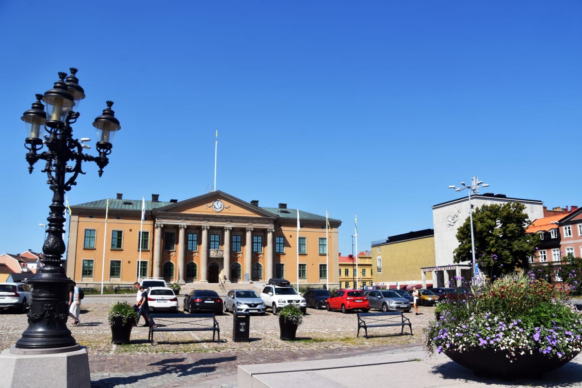 En översiktsbild av ett stadstorg med en stor ljusgul byggnad med pelare och klocka i bakgrunden. Ett antal bilar är parkerade på kullerstenstorget. Två lyktstolpar, blomsterarrangemang och några personer syns på torget under en klarblå himmel.