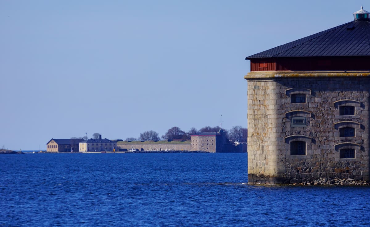 En stor stenbyggnad med mörkt tak står delvis i blått vatten till höger i bilden. I bakgrunden syns en landtunga med flera byggnader, en mur och trädrader utan löv under en klarblå himmel.
