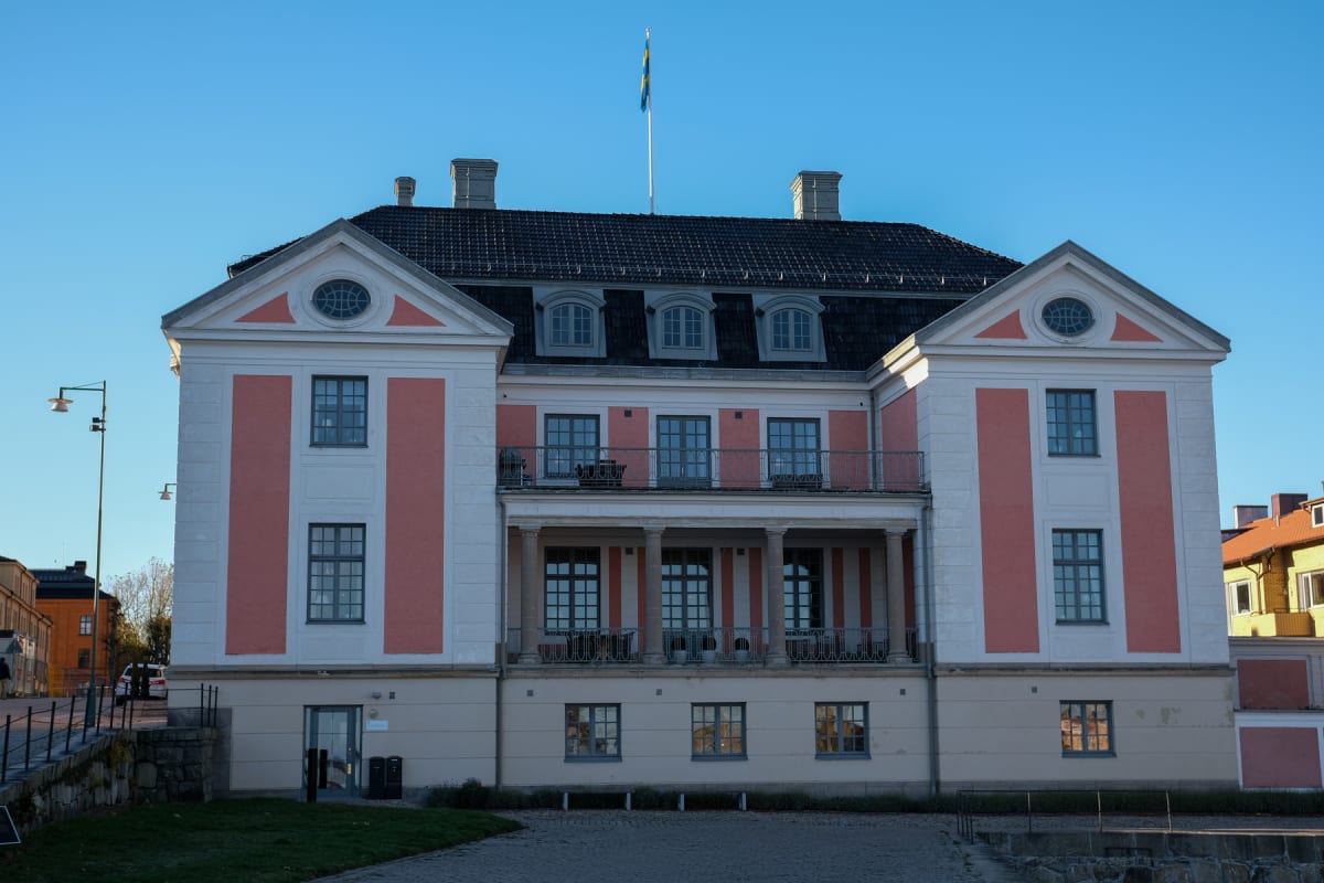 En stor, ljus byggnad med en klassisk fasad i vitt och laxrosa, svart tak och flera fönster. Svenska flaggan syns i topp på en flaggstång på taket. Marken framför byggnaden består av kullersten och gräs, och himlen är klarblå.