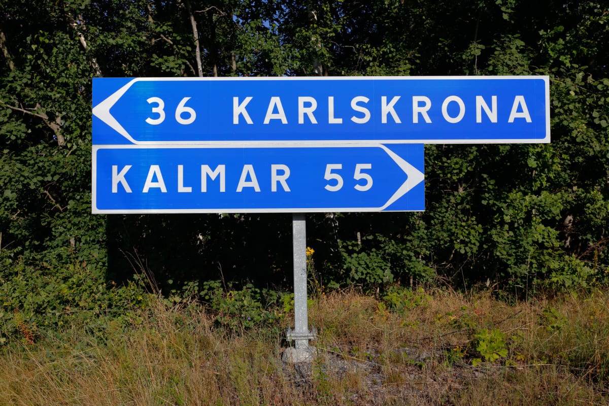 En blå vägskylt med vit text och pilar visar "36 KARLSKRONA" till vänster och "KALMAR 55" till höger. Skylten är monterad på en grå stolpe som står i torrt gräs. I bakgrunden syns tätt, grönt lövverk från träd och buskar under en ljus himmel.