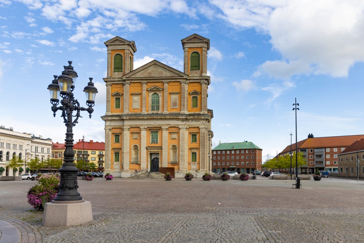 En stor gul byggnad med två torn och kolumner syns i bakgrunden av ett stenbelagt torg. I förgrunden till vänster står en svart utsmyckad gatlykta omgiven av lila blomsterarrangemang. Himlen är blå med vita moln och träden har gröna löv.
