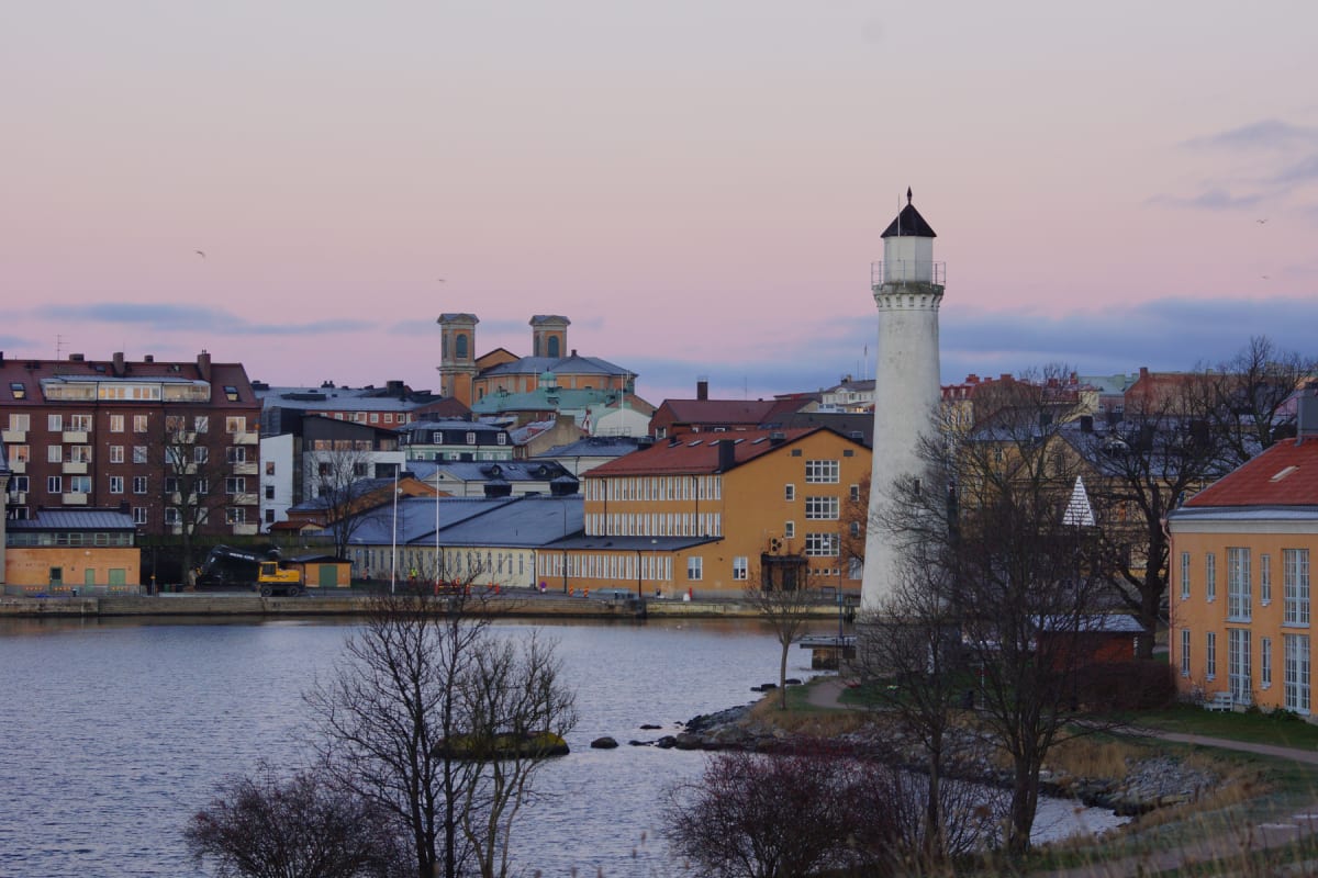 En bild som visar en stadsutsikt med vatten i förgrunden och byggnader samt en fyr i bakgrunden under en gryning eller skymning med rosa-lila himmel. Träd utan löv står längs strandlinjen, och en grävmaskin syns vid vattnet.