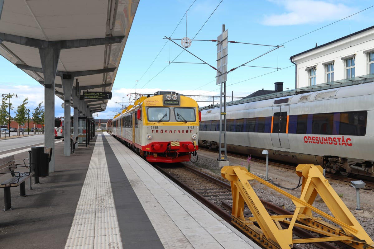 En bild visar ett tåg av typen Krösatåg med gul, röd och vit färgsättning, samt ett Öresundståg i silver och blått, vid en tågperrong under en klarblå himmel. Perrongen har en takkonstruktion, bänkar, en soptunna och en digital informationstavla.