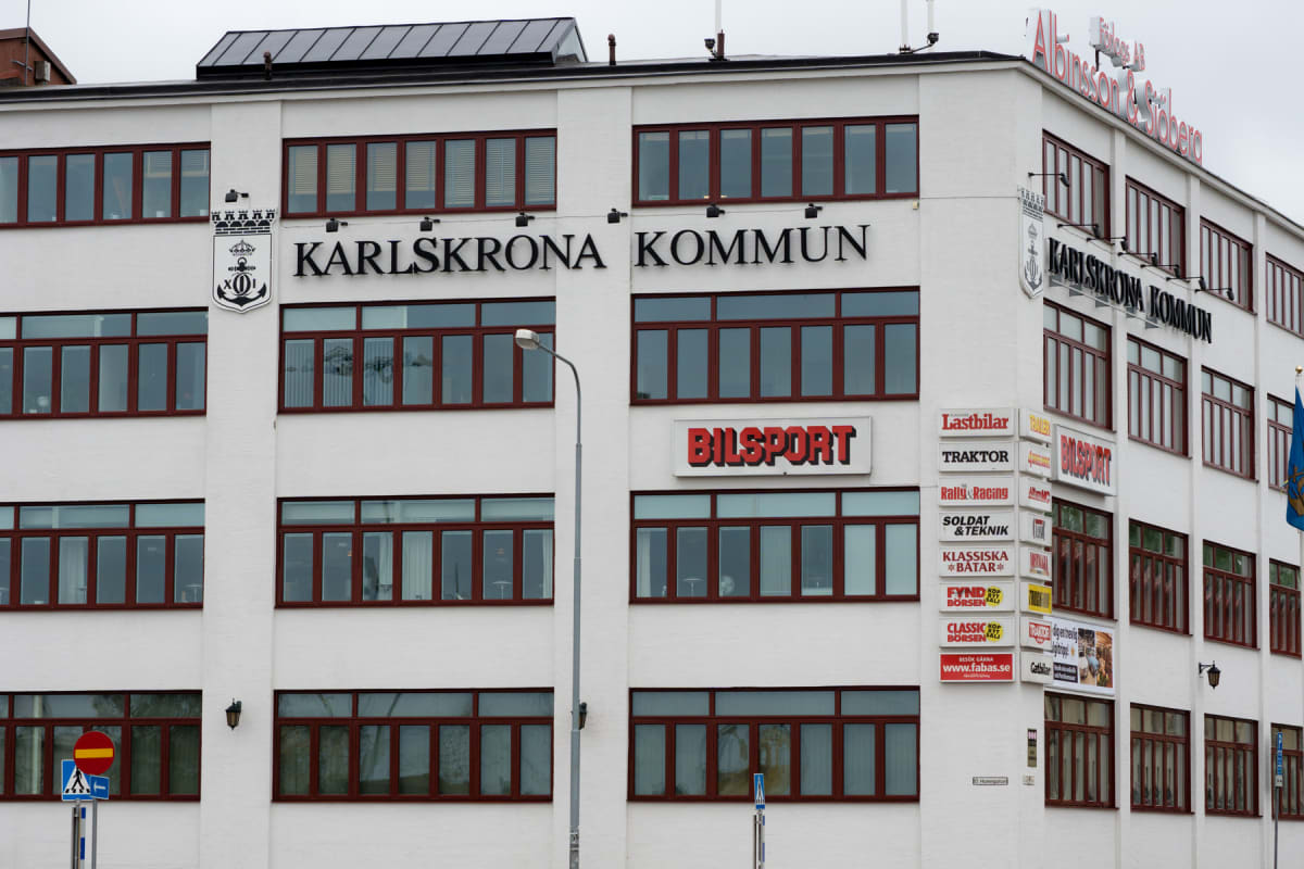 En vit flerplansbyggnad med många bruna fönster syns under en gråmulen himmel. På fasaden finns texten "KARLSKRONA KOMMUN" samt "BILSPORT" och flera skyltar med text som "Lastbilar" och "Traktor". Ett kommunvapen och en gatlykta är också synliga.