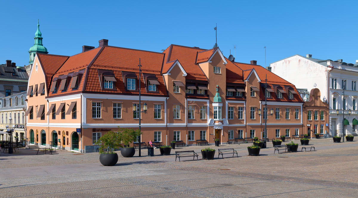 En bild som visar flera äldre byggnader med röda tegeltak och fasader i olika ljusa nyanser, från aprikos till vitt, under en klarblå himmel. Framför byggnaderna finns ett torg med gatsten, prytt med svarta bänkar och planteringskärl med gröna växter