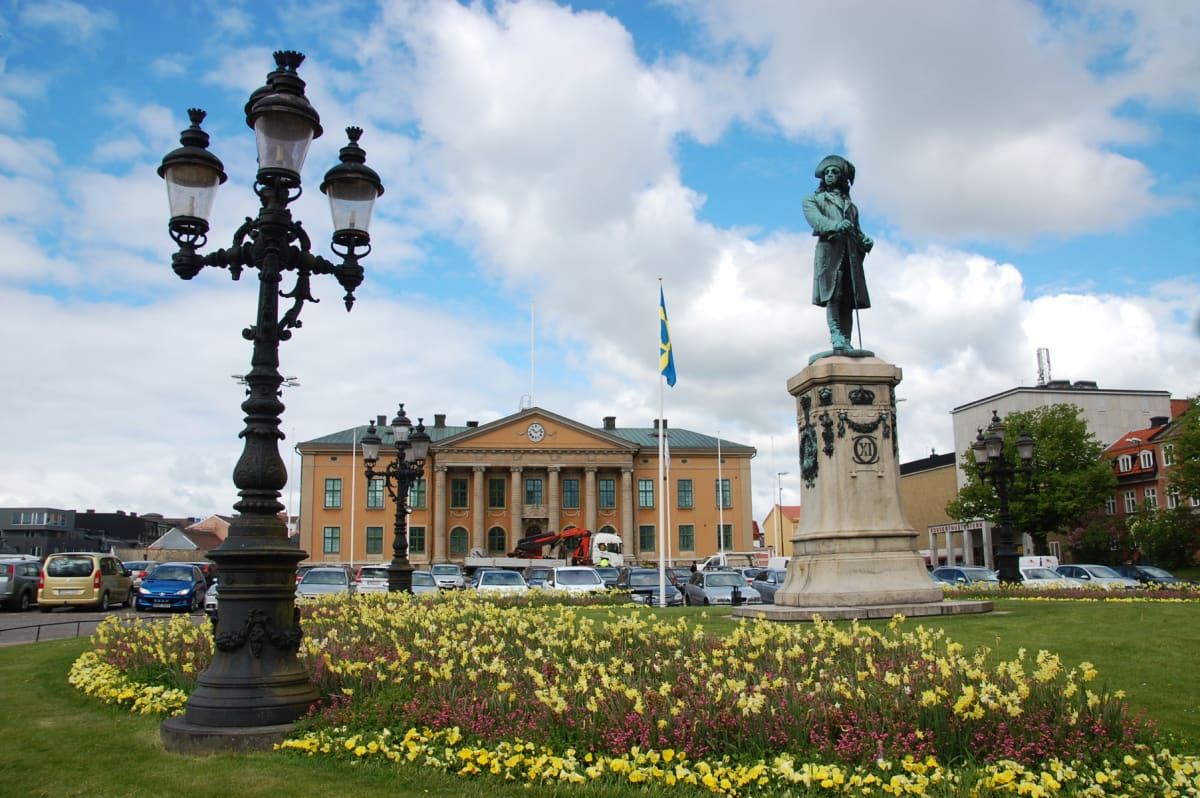 En bild som visar en parkmiljö med en staty och en stor lyktstolpe i förgrunden, omgiven av gräs och gula samt lila blommor. I bakgrunden syns en stor gul byggnad med pelare och klocka, en svensk flagga samt parkerade bilar och ytterligare byggnader
