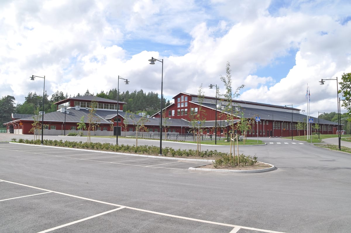En stor, röd byggnad med svart tak och flera fönster under en molnig himmel. Framför byggnaden finns en asfalterad parkeringsplats med vita linjer, gröna planteringar och gatlyktor. I bakgrunden syns tallar.