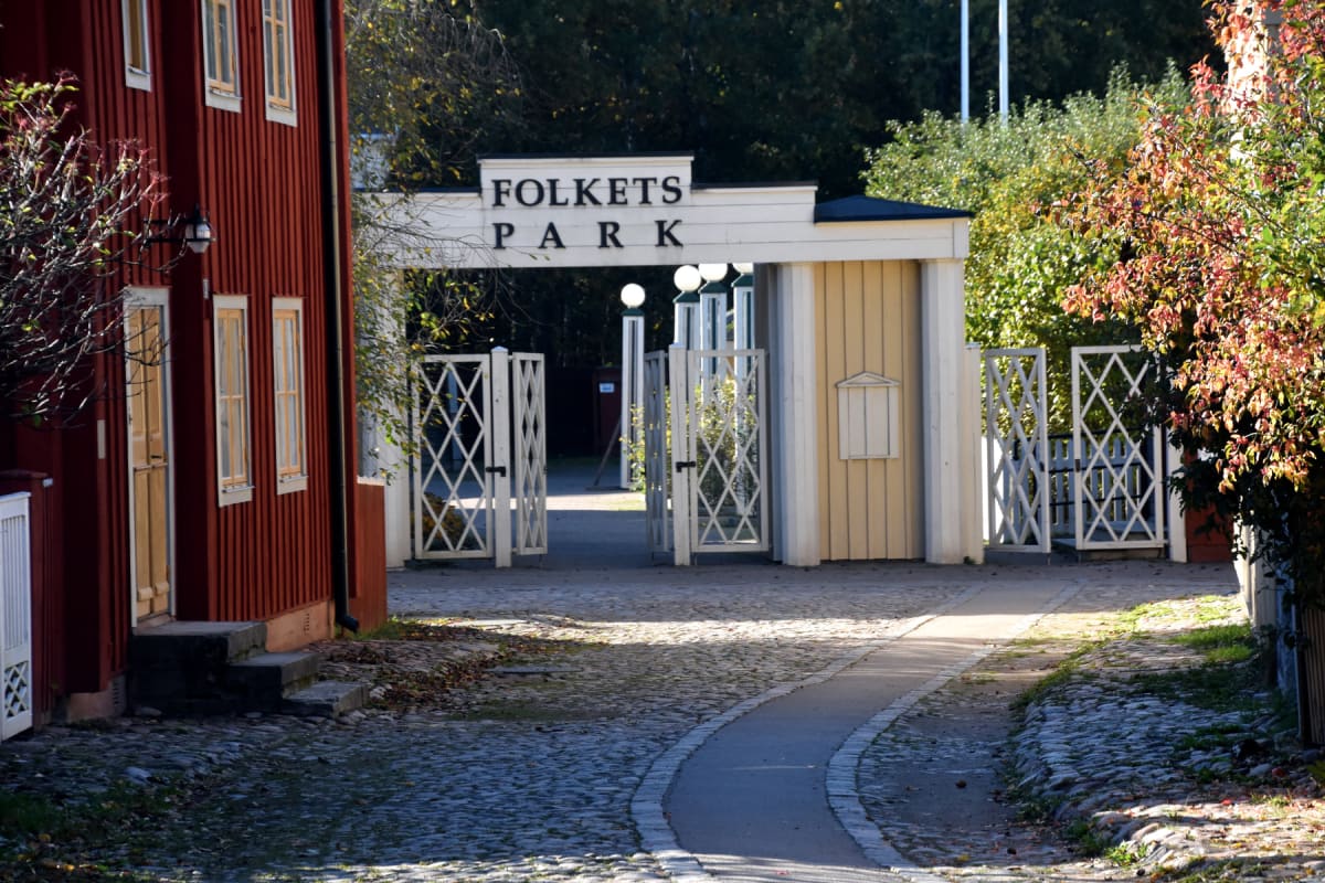En bild av en ingång till Folkets Park med en röd träbyggnad till vänster och gula byggnadsdelar som ramar in skylten. En kullerstensgång med en smal asfaltsgång leder fram till de öppna vita grindarna. Höstlöv syns på marken och på träd.
