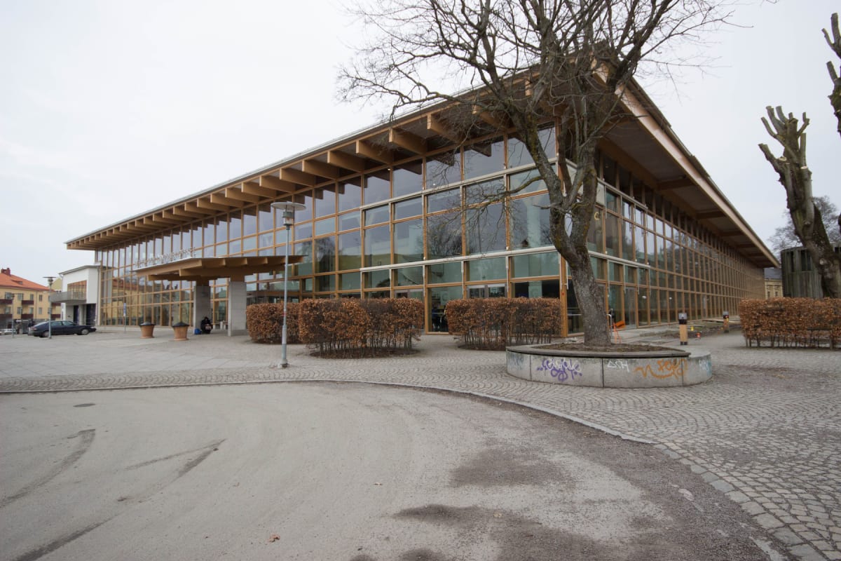 En stor modern byggnad med en fasad av glas och träbalkar framför en öppen plats. Flera träd med kala grenar och buskar med torra löv syns. En person sitter vid byggnadens entré. En mörk bil står parkerad till vänster.