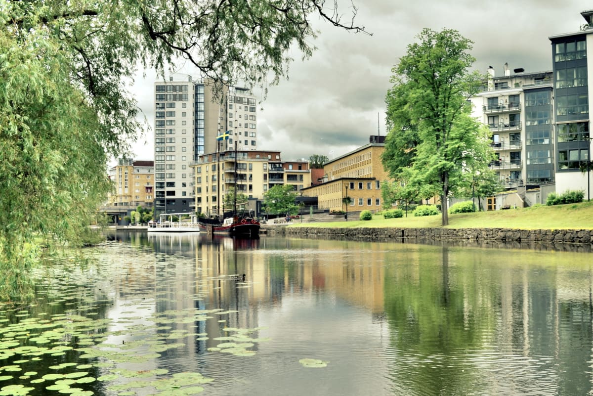 En bild visar en kanal eller flod med byggnader, träd och ett fartyg längs stranden under en molnig himmel. Vattnet reflekterar omgivningen och har näckrosblad. Ett grönt träd och ett segelfartyg syns på höger sida, med en svensk flagga på en av bygg