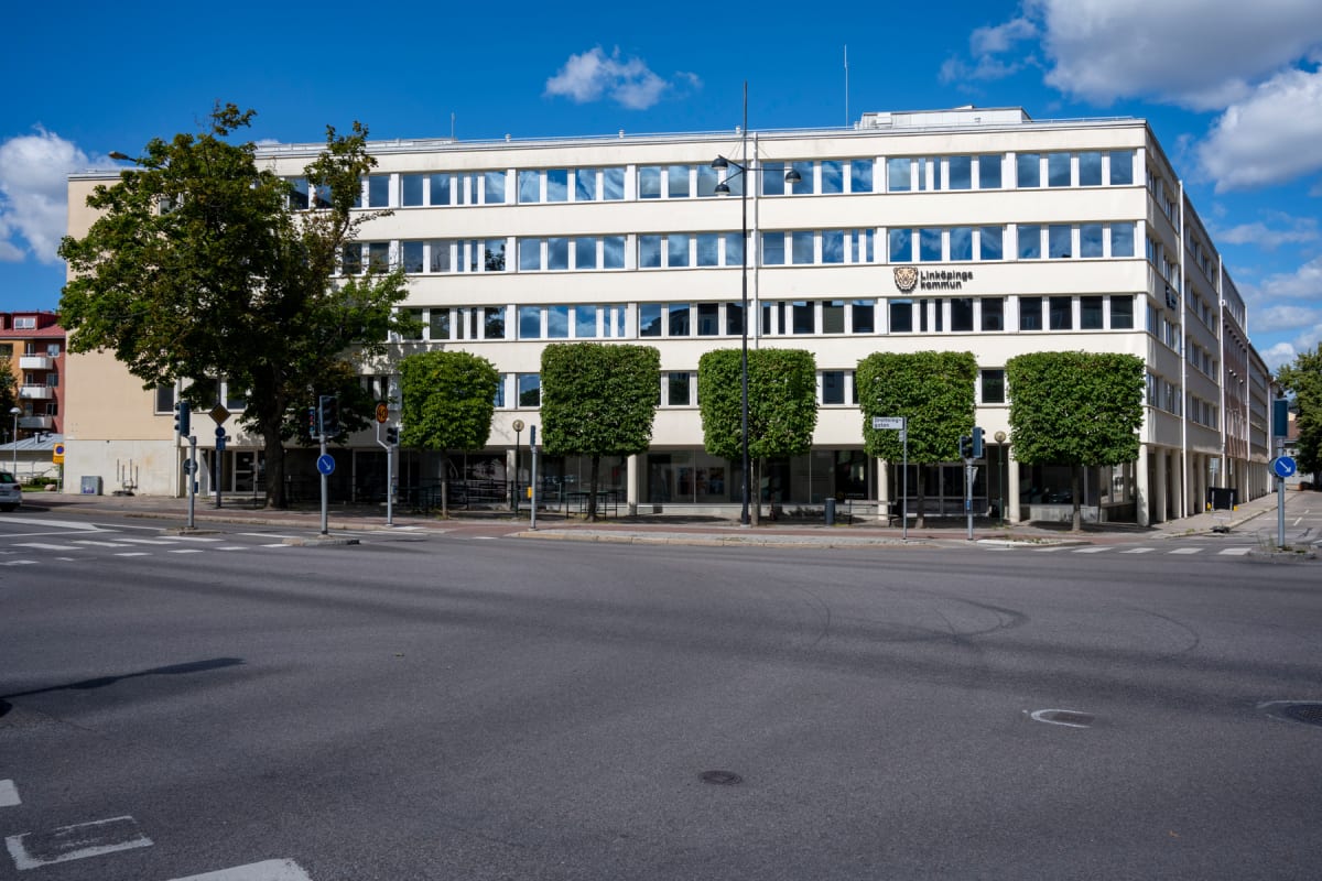 En ljus byggnad med flera våningar och många fönster, med texten "Linköpings kommun" och en logotyp på fasaden. Framför byggnaden finns en gata med övergångsställen, trafikljus, vägskyltar och flera träd, varav några är formklippta. Blå himmel med vi