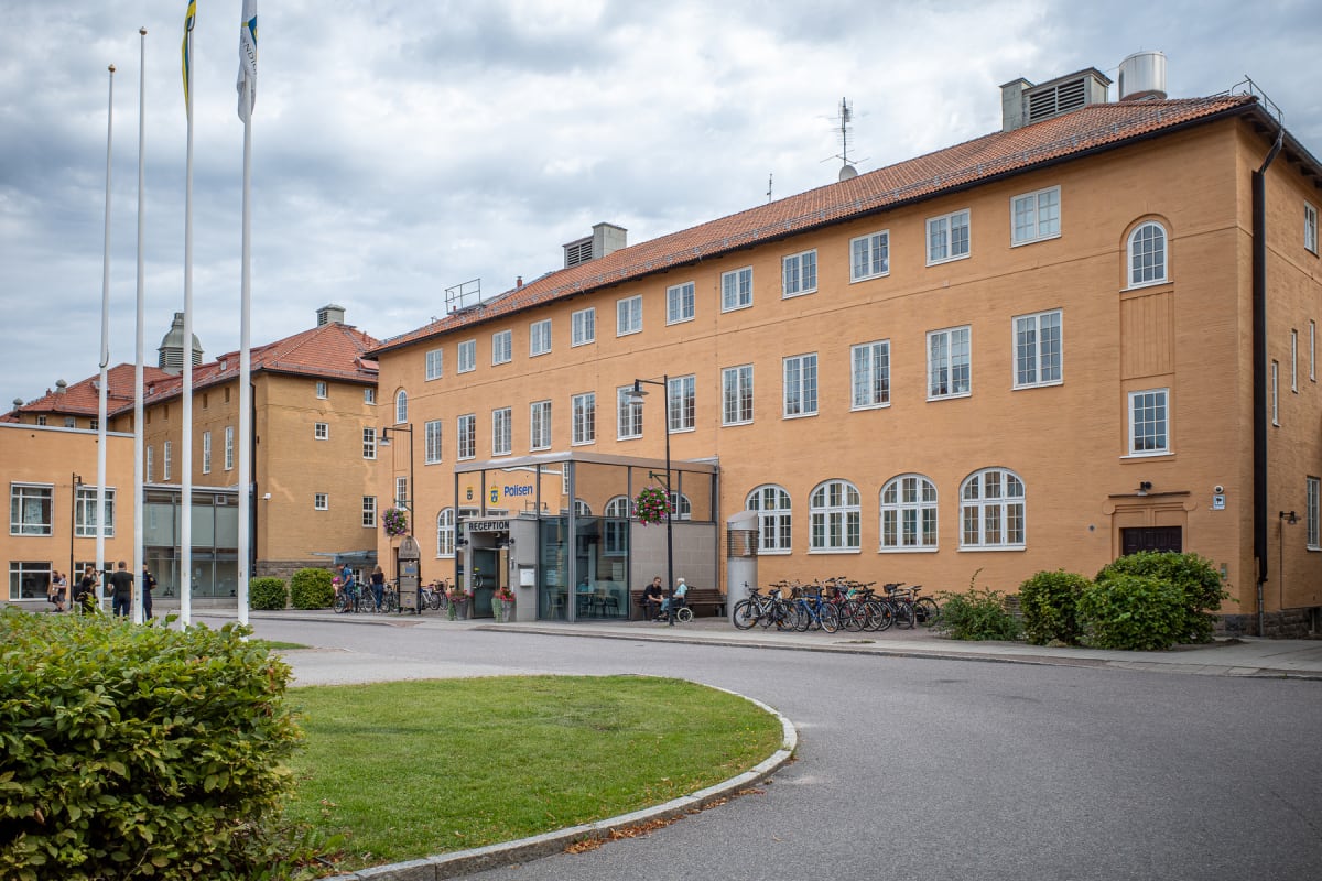 En gata framför flera flerplansbyggnader med orange fasader och röda tegeltak. En av byggnaderna har en skylt märkt "Polisen" och "Reception" vid entrén. Flera cyklar är parkerade utanför och personer ses i närheten. Gräsytor och buskar finns i förgr