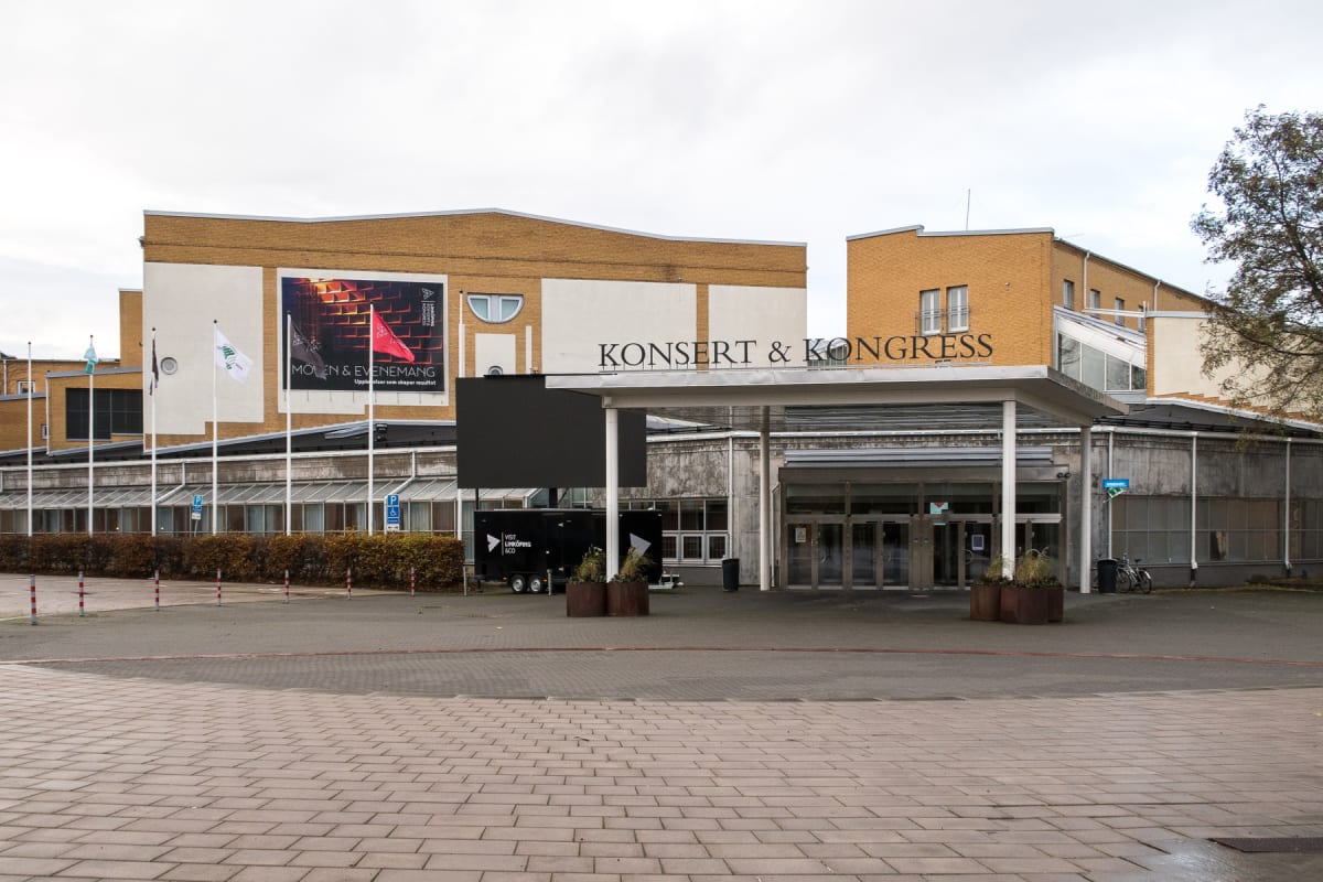 En stor byggnad med gul tegel och vita fasadpartier med texten "KONSERT & KONGRESS" ovanför entrén. Framför byggnaden finns ett stort markområde med gatsten och asfalterade ytor, några flaggstänger med flaggor samt en svart släpvagn med en stor LED-s