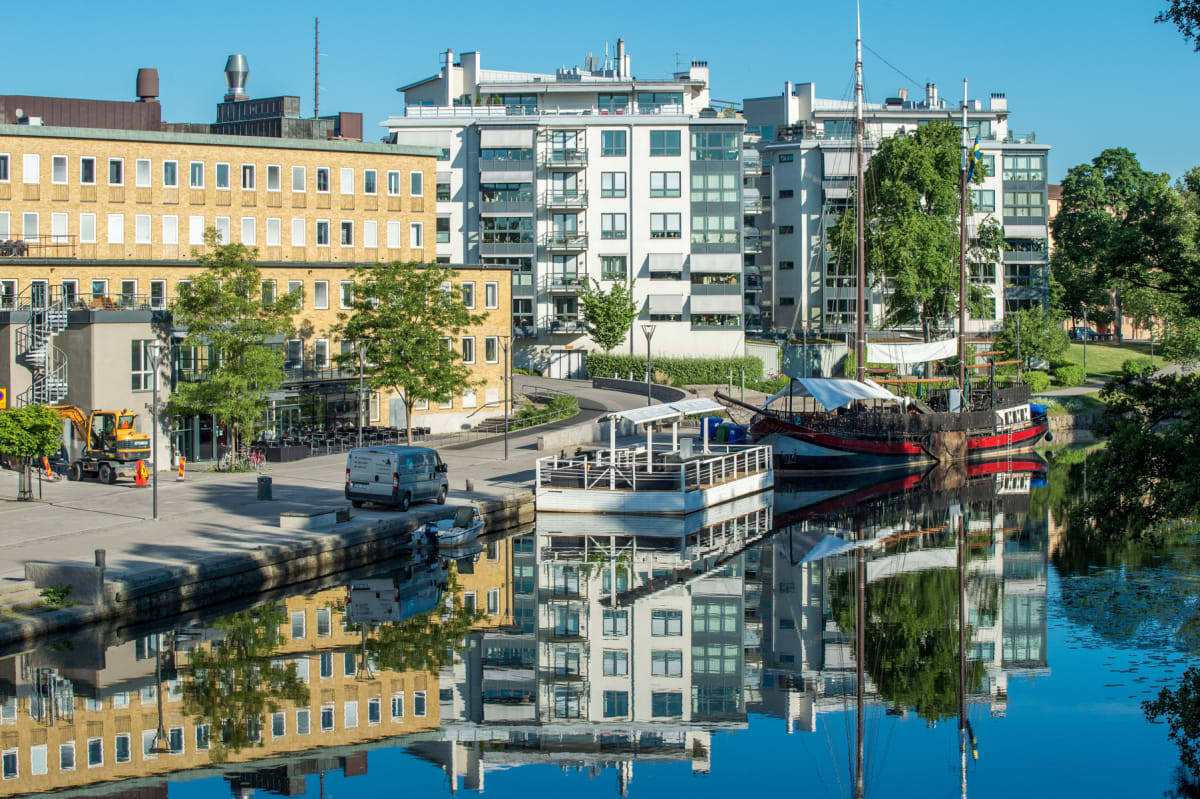 En översiktsbild som visar byggnader, en kanal med båtar, grönska och fordon under en solig dag. Ett gult tegelhus och flera moderna vita byggnader speglar sig i vattnet bredvid en större segelbåt och en vit flytbrygga. En grävmaskin och en skåpbil s