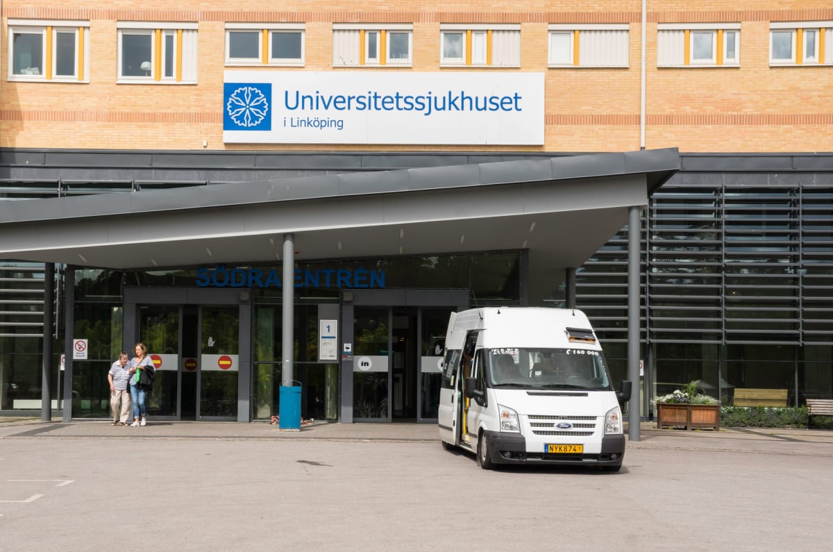 En vit minibuss av märket Ford står parkerad framför "Södra entrén" till Universitetssjukhuset i Linköping. Byggnaden är i tegel med stora fönsterpartier. Två personer går vid entrén. En skylt med "Universitetssjukhuset i Linköping" syns.