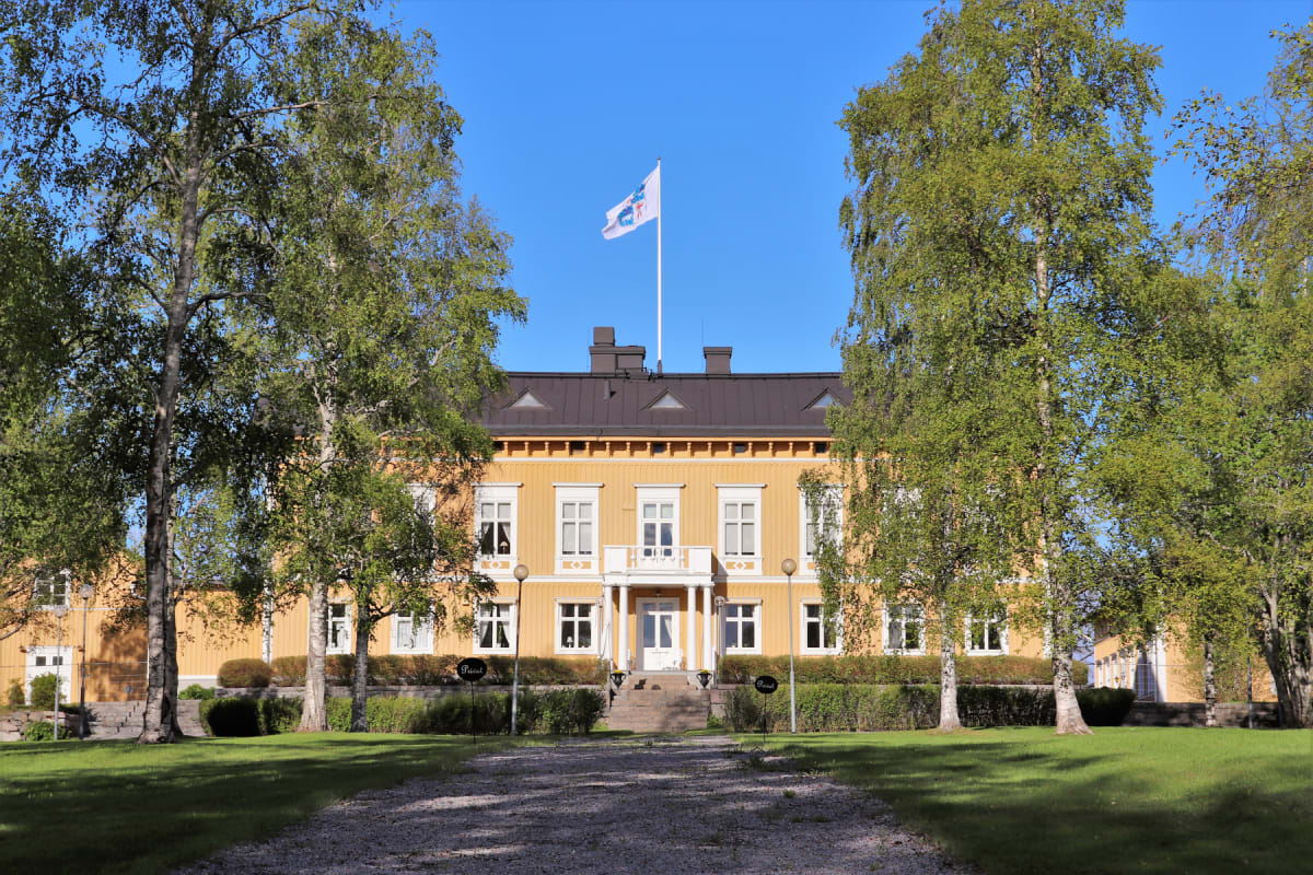 En stor, gul byggnad med vita fönsterkarmar och ett mörkt tak står centralt i bilden. Framför byggnaden finns en grusgång som leder upp till en trappa och en balkong. Två stora lövträd med gröna blad ramar in byggnaden. En flaggstång med en vit flagg