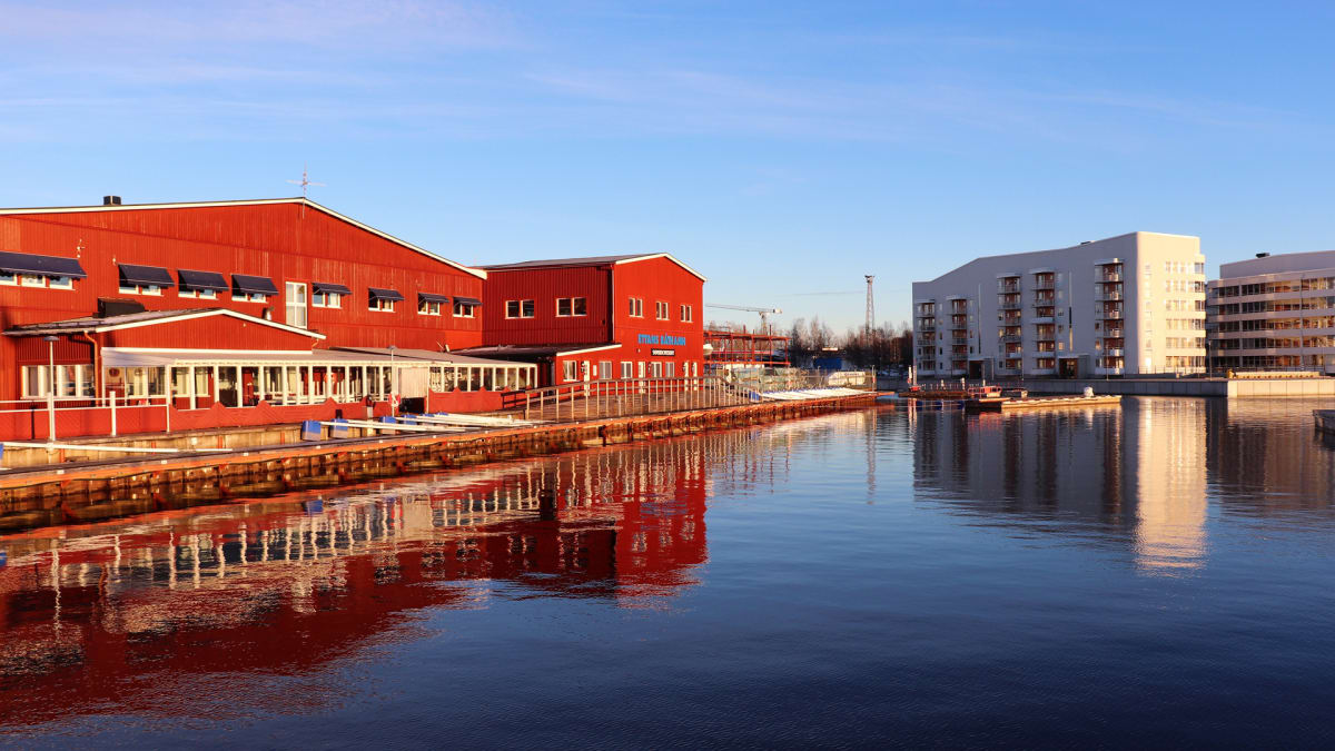 En bild som visar en stillsam vattenyta med byggnader på båda sidor. Till vänster syns flera röda träbyggnader med vita fönster och mörka markiser, delvis reflekterade i vattnet. Till höger syns moderna flerbostadshus med balkonger, även de reflekter