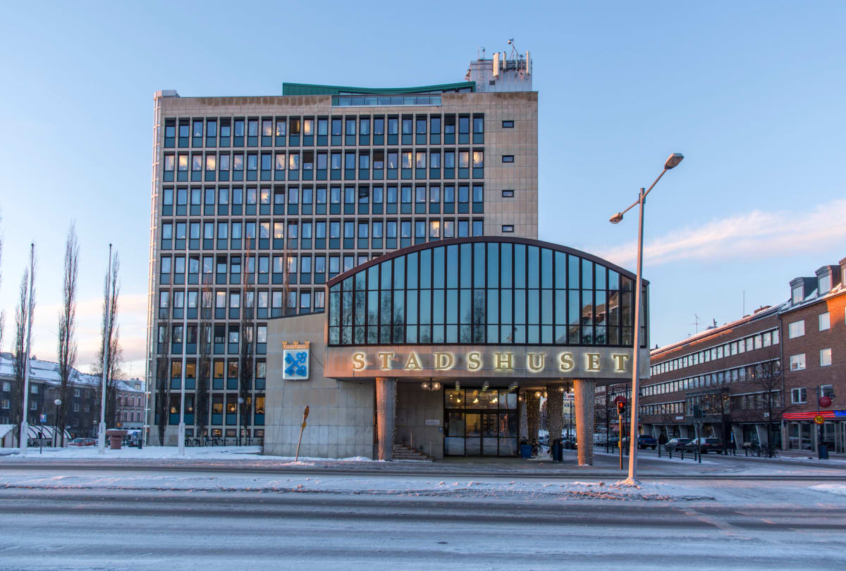 En fasadvy av en stor byggnad med texten "STADSHUSET" upplyst över entrén, omgiven av mindre byggnader och en snötäckt gata. Byggnaden har många fönster och en logotyp bredvid entrén. Det är dagsljus men belysningen vid entrén är tänd.