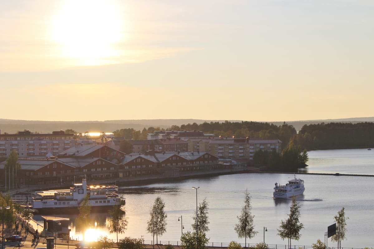 En översiktsbild av en stad vid vattenlinjen under solnedgång eller soluppgång. Solen står högt i bild och reflekteras i vattnet. Flera byggnader, träd och två båtar syns. En båt ligger vid kaj, en annan färdas på vattnet.