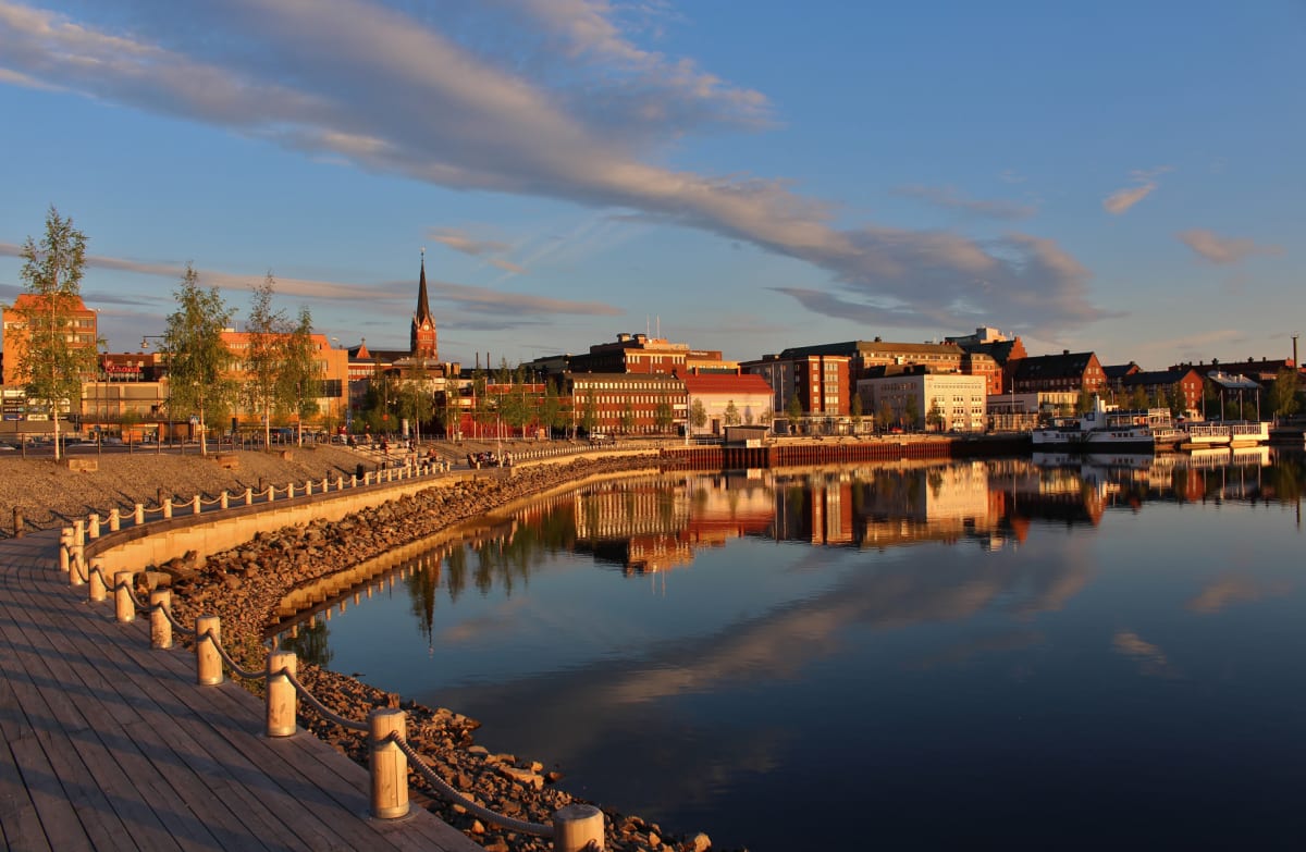 En vidvinkelbild av en stadssilhuett vid en lugn vattenyta under en solnedgång eller soluppgång. Byggnader med mestadels röda och orange takfasader speglas i vattnet, tillsammans med en kyrkspira. En träbrygga med rep följer strandlinjen.