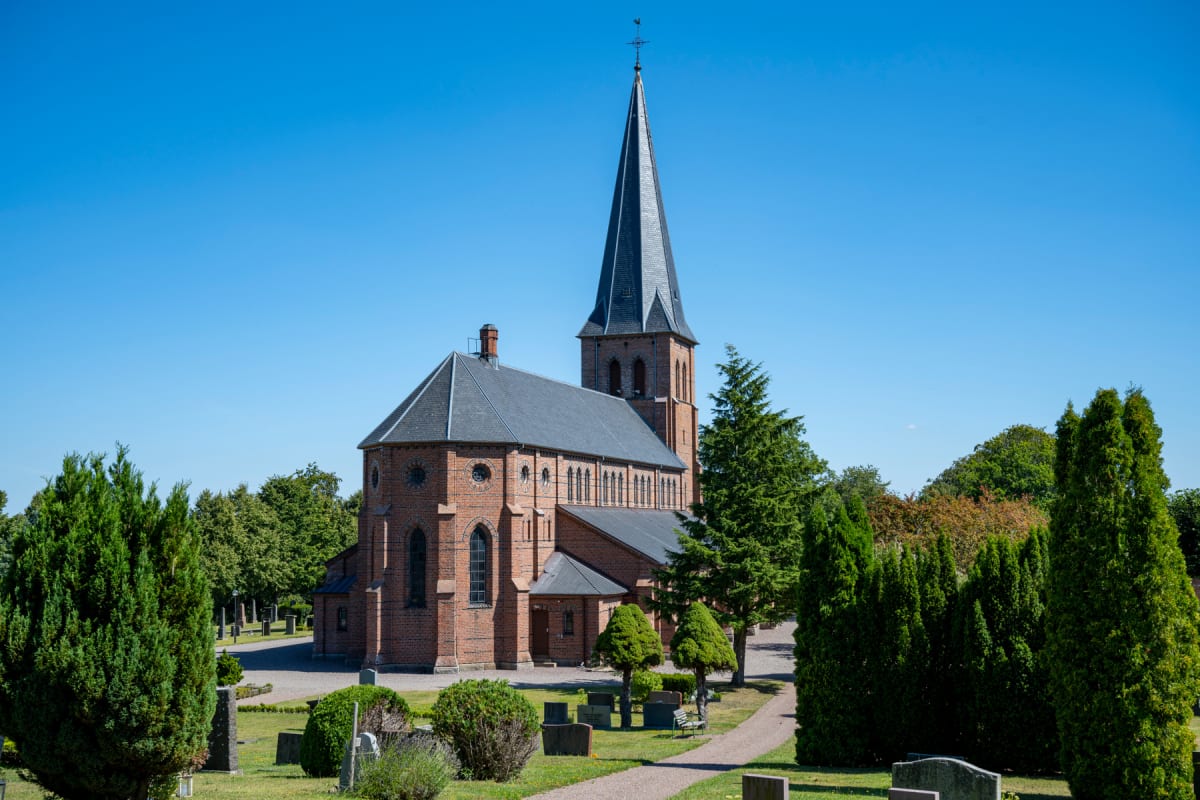 En röd tegelkyrka med mörkt tak och en hög spira står under en klarblå himmel. Runt kyrkan syns en kyrkogård med gräs, grusgångar, olika träd, buskar och gravstenar. Bilden är tagen utomhus i dagsljus.