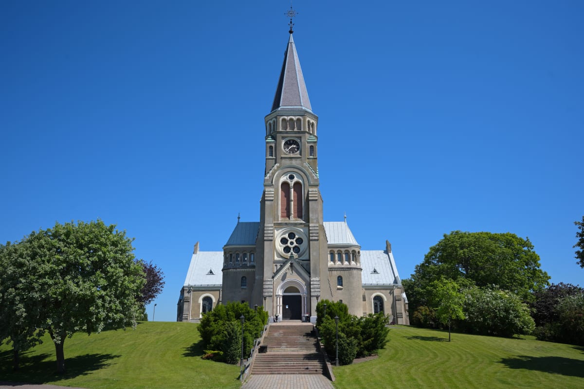 En stor, ljus kyrkbyggnad med en hög spira och klocktorn syns framifrån under klarblå himmel. Framför kyrkan leder en stenlagd trappa uppför en gräsklädd sluttning. Träd och buskar kantar sidorna av kyrkan och gräsmattan.