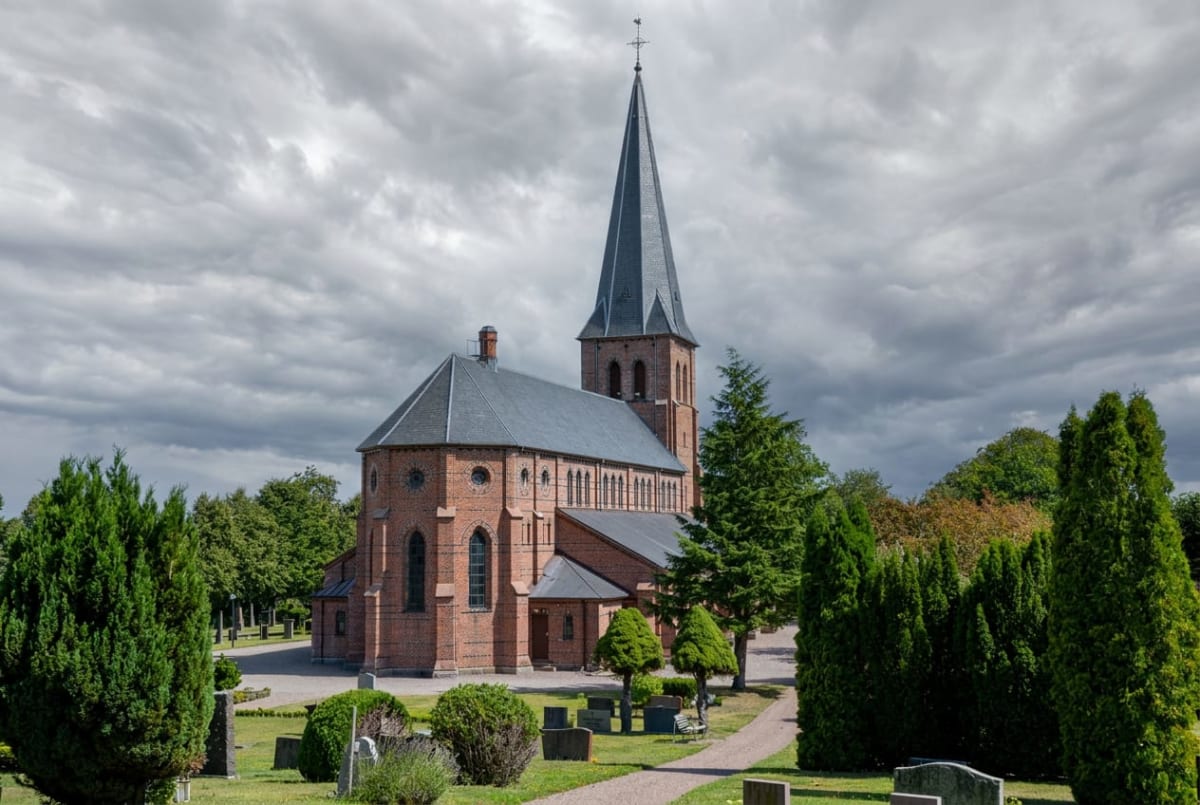 En röd tegelkyrka med ett grått tak och en spetsig spira står på en gräsklädd kyrkogård. Runt kyrkan syns flera träd i olika gröna nyanser och gravstenar. Himlen är delvis molnig.