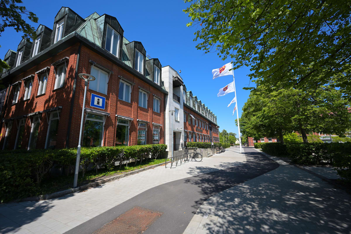 En röd tegelbyggnad med grönt plåttak under en klarblå himmel. Flera gröna träd skuggar en gångväg intill byggnaden. Utanför byggnaden finns tre vita flaggor med ett rött vapen, en lyktstolpe med en informationsskylt och en cykel parkerad vid ett cyk