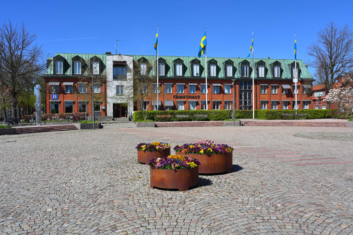 En fasadvy av en röd tegelsbyggnad med ett grönt tak och flera svenska flaggor vajande, framför en kullerstensbelagd torgplats. Tre blomsterurnor med färgglada blommor står i förgrunden, och på byggnaden syns texten "KOMMUNHUS" samt en informationssk