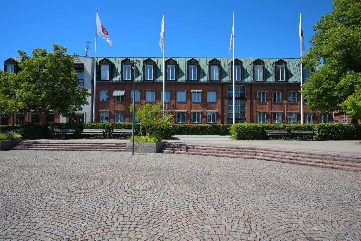 En röd tegelbyggnad med grönmålade tak och vita fönsterkarmar under en klarblå himmel. Framför byggnaden finns en stensatt yta, trappsteg, gröna buskar, träd och tre flaggstänger, varav två bär vita flaggor med symboler.