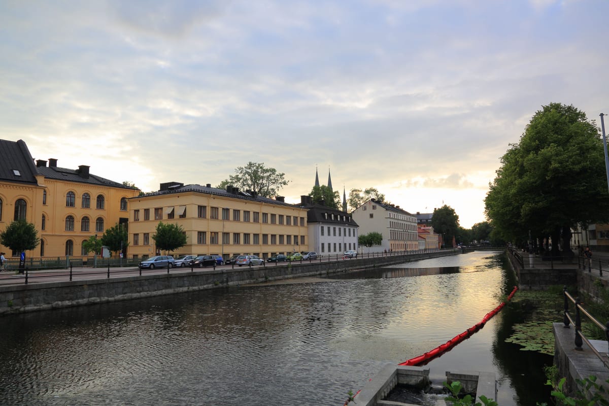 En bild som visar en stadskanal med byggnader och träd längs sidorna, med bilar parkerade vid kajen på vänster sida. Två spiror från en byggnad syns i bakgrunden. Ett rött flytblock ligger i vattnet till höger. Himlen är molnig med ljusare partier.