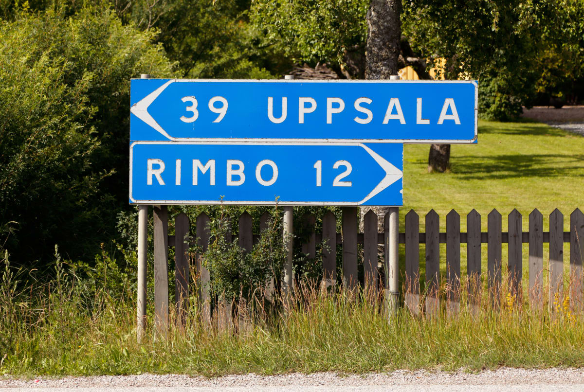 En blå vägskylt med vita texter och pilar står vid sidan av en grusväg. Skylten pekar mot "39 UPPSALA" till vänster och "RIMBO 12" till höger. I bakgrunden syns grönskande träd och buskar, samt en trästaket och gräs.