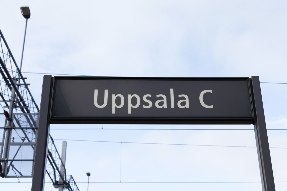 En svart skylt med vit text "Uppsala C" syns mot en ljusblå himmel med moln. Till vänster och i bakgrunden syns delar av en metallkonstruktion, som liknar järnvägsinfrastruktur, med strömkablar och en gatlykta.