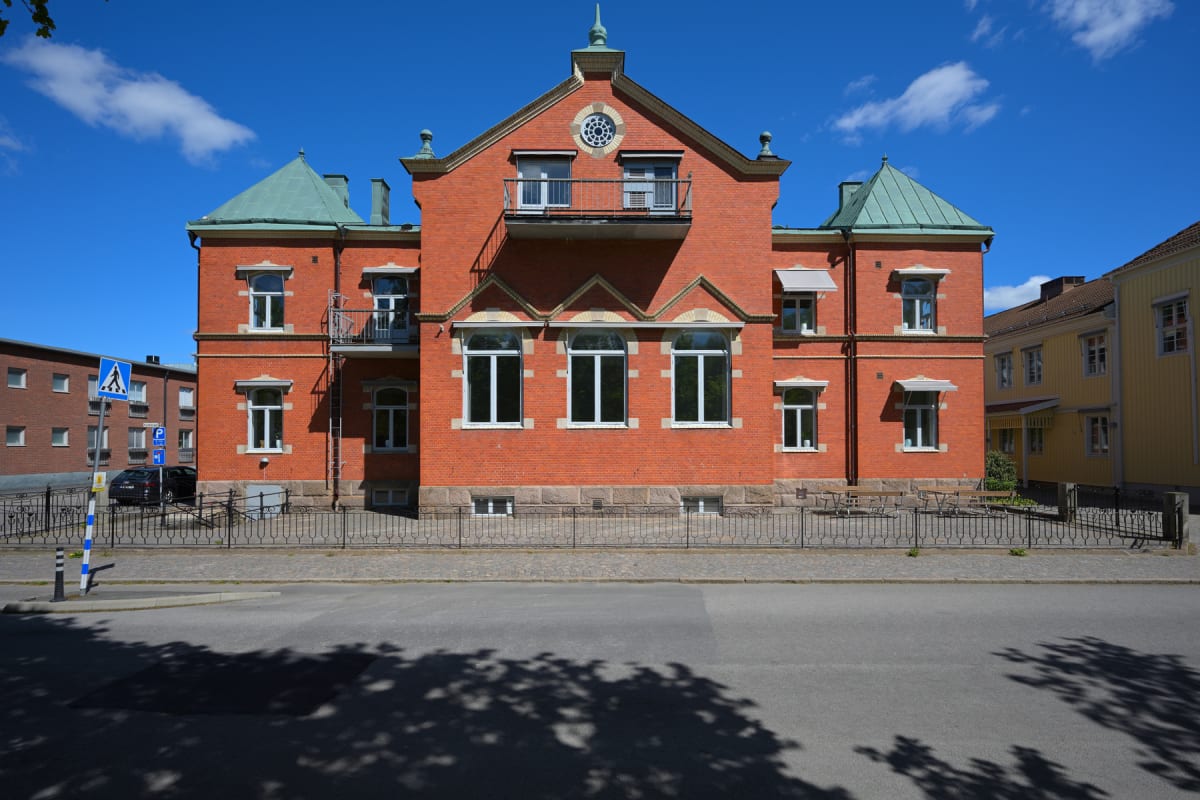 En röd tegelbyggnad med gröna tak, vita fönster och balkonger, syns från gatan under en klarblå himmel med några moln. Framför byggnaden finns ett svart smidesstaket, en trottoar med gatsten och en asfalterad väg med trädskuggor.