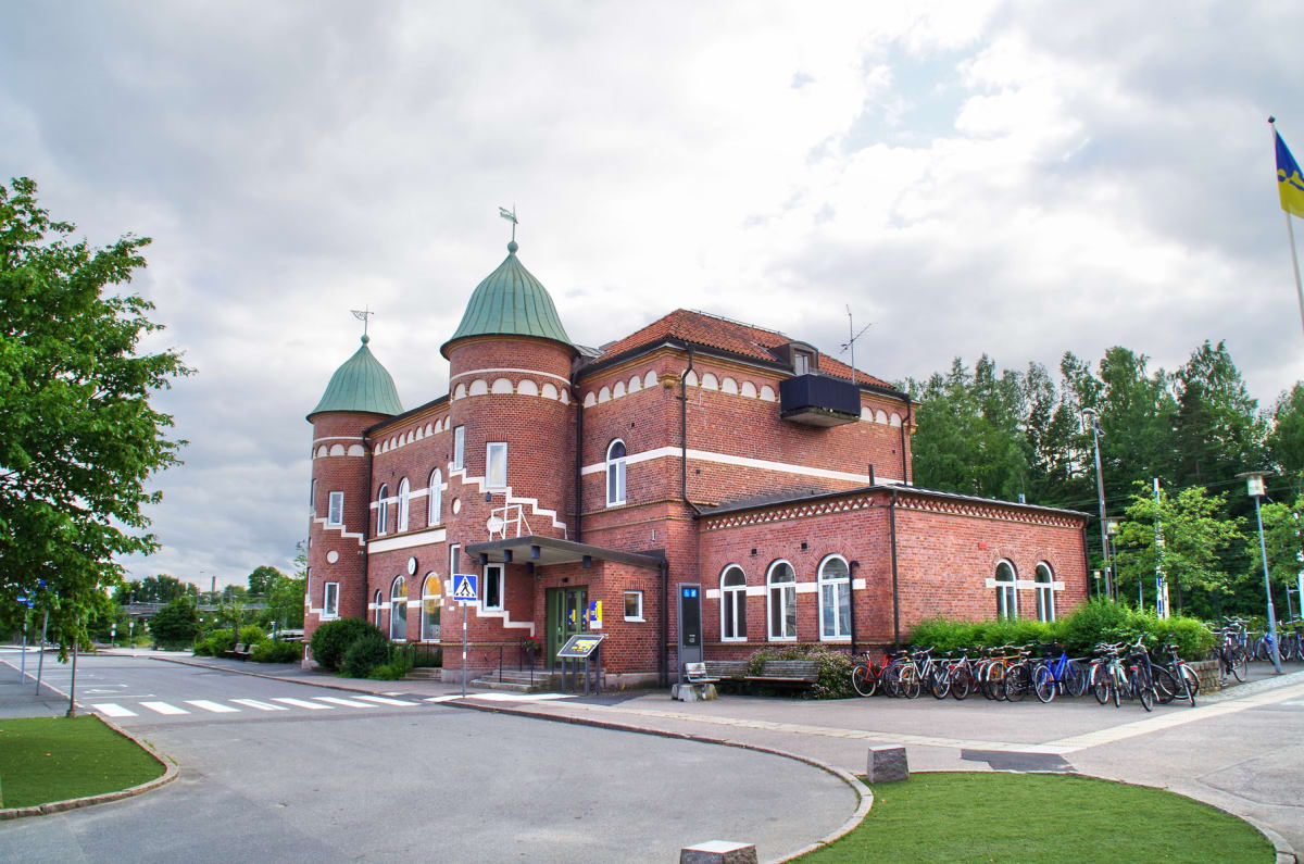 En röd tegelbyggnad med två grönkupiga torn står vid en asfalterad väg med ett övergångsställe. Träd med gröna löv och cykelställ med flera cyklar syns. En flaggstång med en gul och blå flagga är synlig till höger, och himlen är molnig.