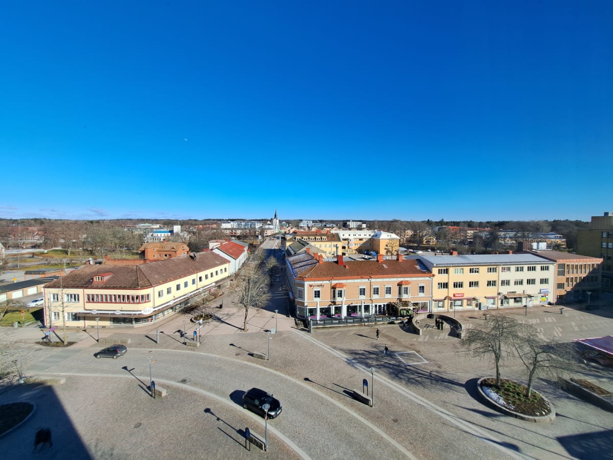 Bilden visar en översikt över en stadskärna med en torgplats och en gata kantad av byggnader under en solig dag. Flera bilar är parkerade eller kör på gatan, och några personer syns på torget och trottoarerna. Himlen är klarblå och inga löv finns på