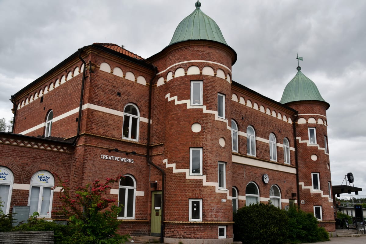 En röd tegelbyggnad med detaljerad arkitektur, inklusive runda torn med grönpatinerade koppartak, vita horisontella band och vita bågformade fält under takfoten. Byggnaden har många vita fönster, och vissa fönster är välvda. Buskar med röda blommor s