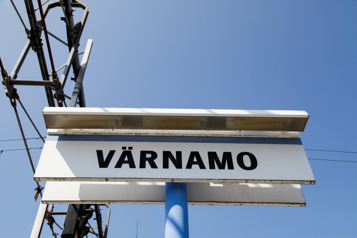 En vit skylt med texten "VÄRNAMO" i svarta versaler, upphängd under en blå stolpe. En metallkonstruktion och kablar syns till vänster i bilden, mot en klarblå himmel. Skugga faller över texten.