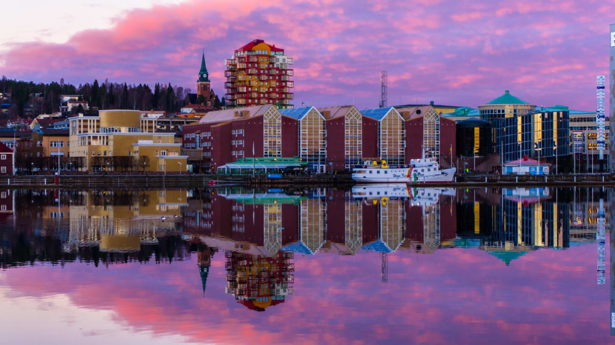 En panoramabild av en stadssiluett vid vatten vid solnedgång eller soluppgång, där byggnader och himlen med rosa och lila moln reflekteras i vattnet. I förgrunden syns ett flertal byggnader, en kyrka med spira, ett färgglatt höghus och en båt förtöjd