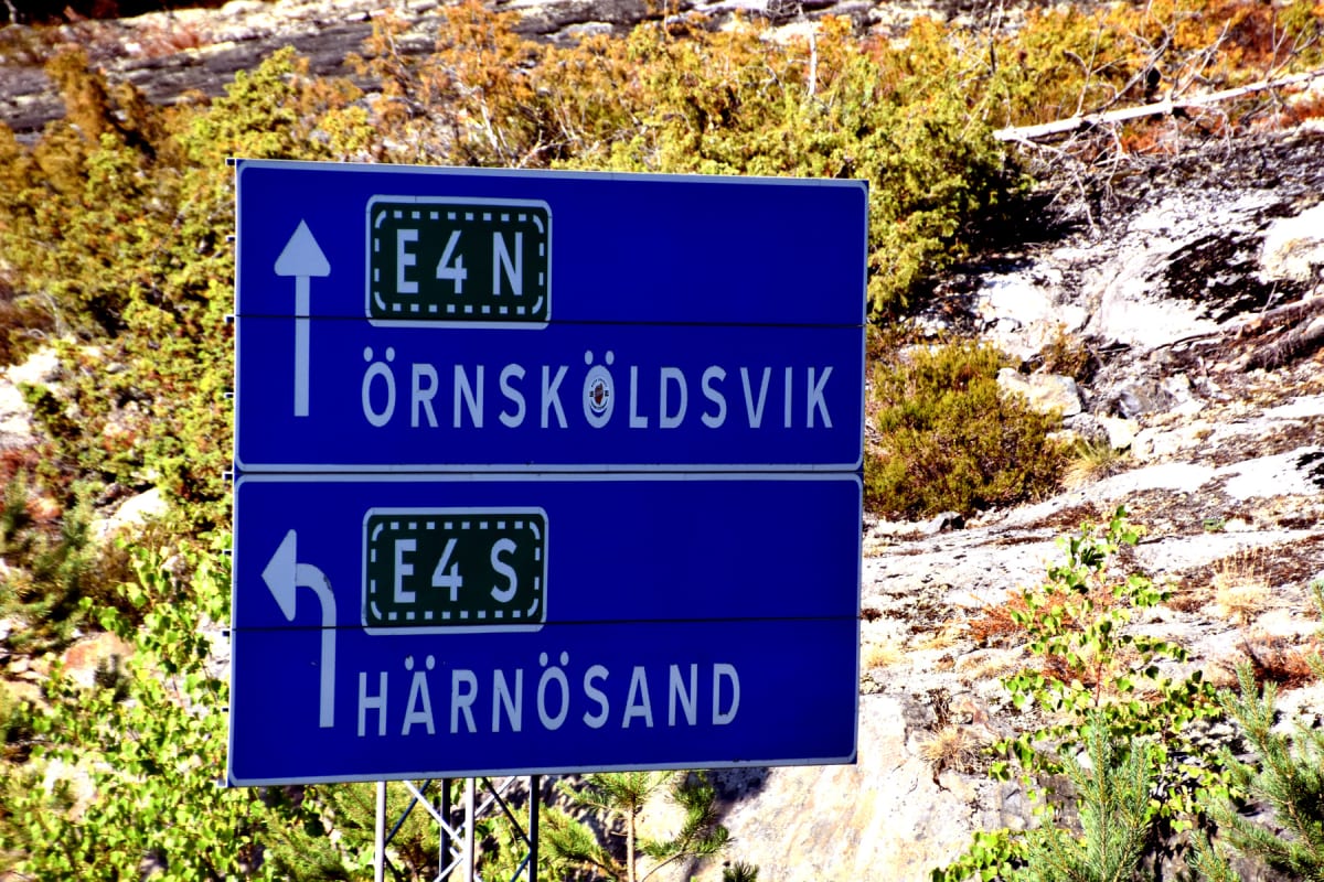 En blå vägskylt med vita pilar och texten "E4 N ÖRNSKÖLDSVIK" och "E4 S HÄRNÖSAND" står vid sidan av en bergig terräng med buskar och träd i bakgrunden. Vegetationens färger indikerar höst.