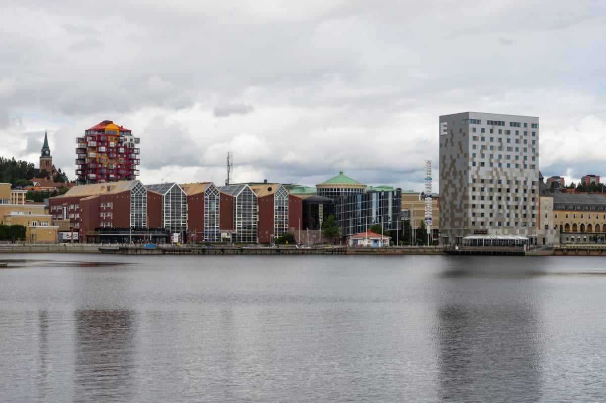 En översiktsbild av en stadssilhuett vid vattnet under molnig himmel. Flera byggnader med olika arkitektur, inklusive ett högt färgglatt hus, en kyrka med spira, och ett grått hotell med 'E' på fasaden, kantar en vattenyta.