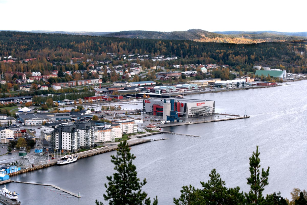 En översiktsbild av en stad vid vattnet, med en stor arena eller byggnad märkt "Fjällräven Center" och "Fjällräven", samt bostadshus, hamnområde med båtar, och skogsklädda berg i bakgrunden. Träd med höstfärger syns tydligt.