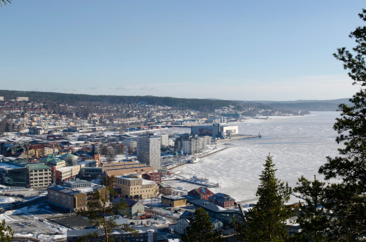 En vid översiktsbild av en stad och en frusen vattenyta under en klarblå himmel. Staden har byggnader av varierande storlek och arkitektur, med snö på marken. Vid vattnet syns en kaj och några mindre fartyg.
