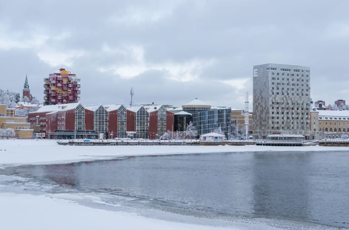 En bild över en stad med flera byggnader längs en delvis isbelagd vattenyta under en molnig vinterdag. Byggnaderna varierar i stil och höjd, inklusive en hög, smal byggnad med mönstrad fasad och en röd- och gulrandig byggnad med balkonger. En kyrktor