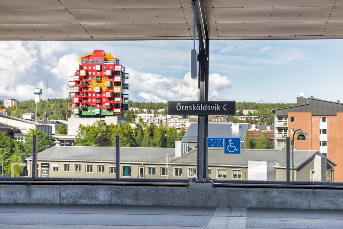 En bild tagen från en tågperrong som visar ett "Örnsköldsvik C"-skylt, delar av perrongtaket och en stadssiluett i bakgrunden. En flerfärgad byggnad i rött, gult och grönt sticker ut mot en himmel med vita moln och blått. Grönska syns på kullar.