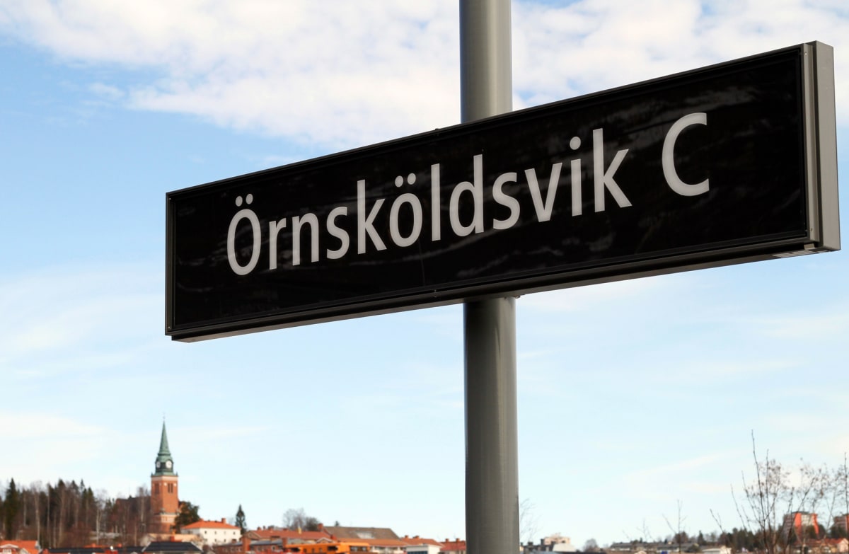 En svart skylt med vit text "Örnsköldsvik C" är monterad på en grå stolpe, mot en bakgrund av en blå himmel med vita moln. I bakgrunden syns taket på en byggnad med en grönspira, träd och bebyggelse.